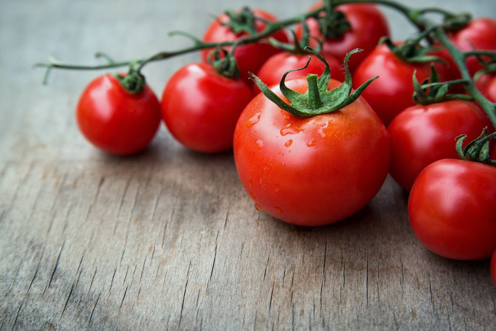 Close-up to red tomatoes attached to a green vine on a wooden table.