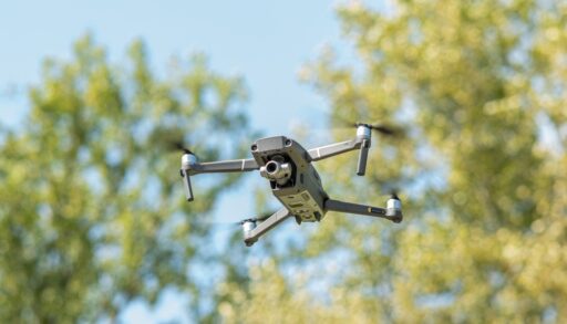 A grey drone with a camera flying in the sky with green trees in the background.