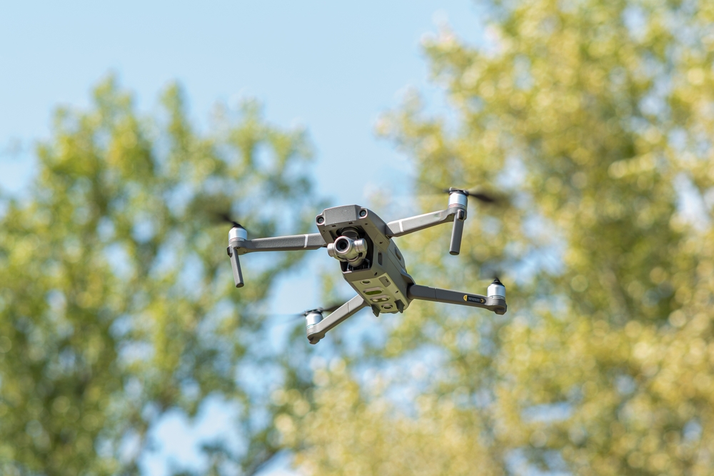 A grey drone with a camera flying in the sky with green trees in the background.