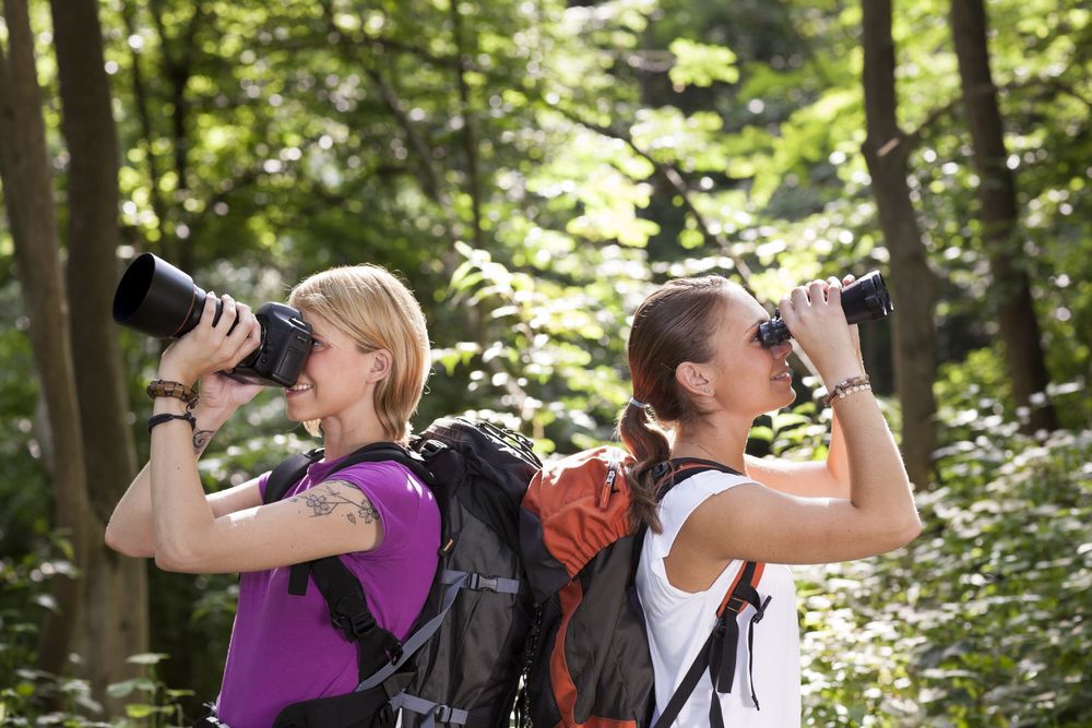 Two women looking through binoculars and a camera while birdwatching.