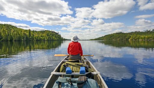 Canoeist sitting in the front of a canoe on a lake with green trees on either side.