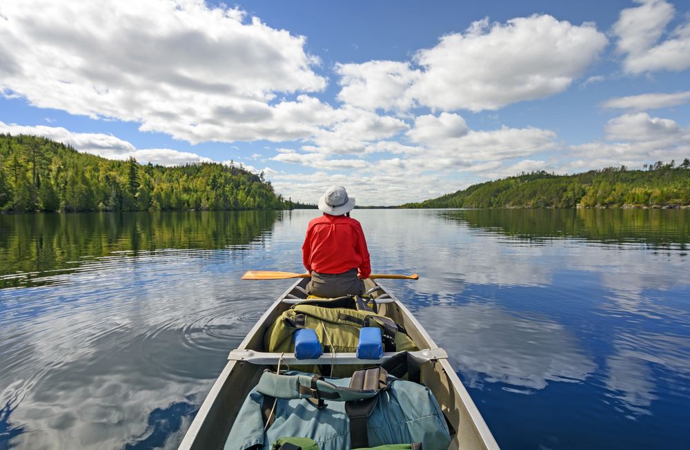 Canoeist sitting in the front of a canoe on a lake with green trees on either side.