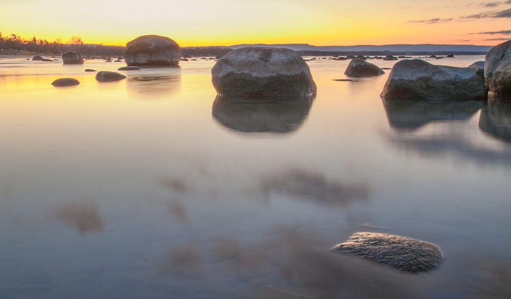 Sunset over a still Lake Huron with large rocks standing in the water.