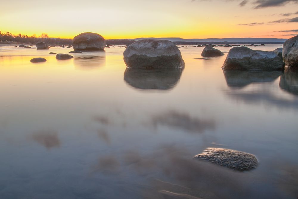 Sunset over a still Lake Huron with large rocks standing in the water.