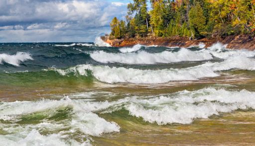 Blue and green waves crashing on the shores of Lake Superior with a green forest surrounding the water.