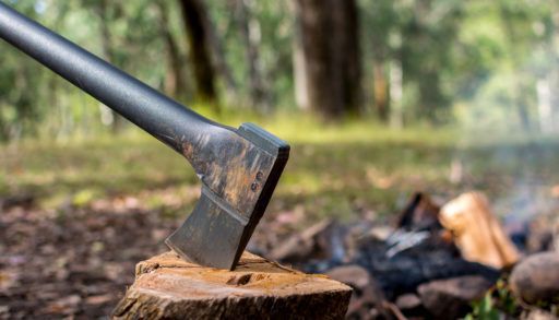 A silver axe stuck in a piece of wood next to a pile of chopped wood in the forest.