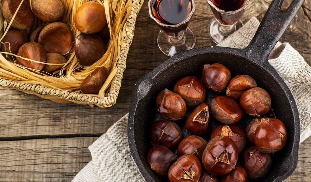 Flat-lay of roasted chestnuts in a cast iron pan next to two drink glasses filled with wine.