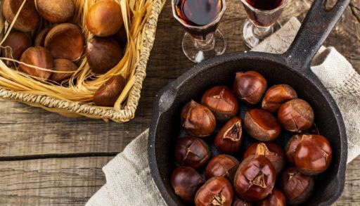 Flat-lay of roasted chestnuts in a cast iron pan next to two drink glasses filled with wine.