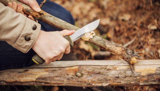 A woman using a hunting knife to cut the bark off a branch.