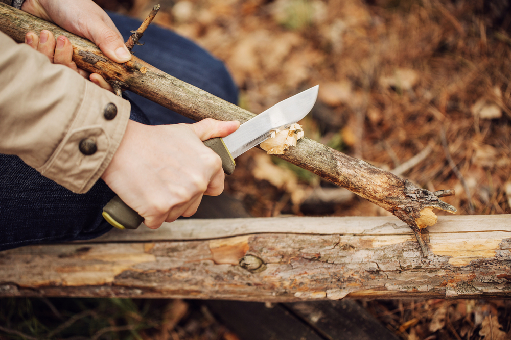 A woman using a hunting knife to cut the bark off a branch.