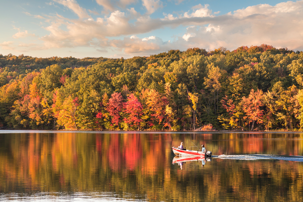 A small, red fishing boat on a glassy lake with the autumn trees reflected in its surface.