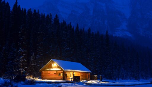 Illuminated log cabin surrounded by snow and trees at dusk in Banff National Park, Alberta.