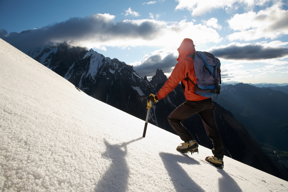 Person hiking up a mountain in an orange jacket with more mountains in the background.
