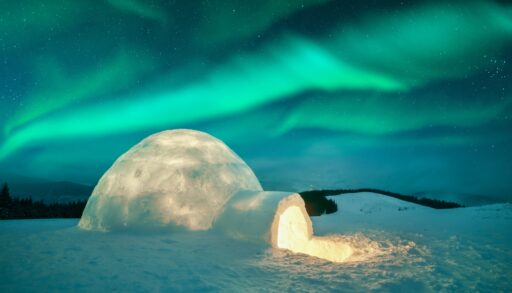 Igloo with a light coming from it against the Northern lights and mountain landscape.