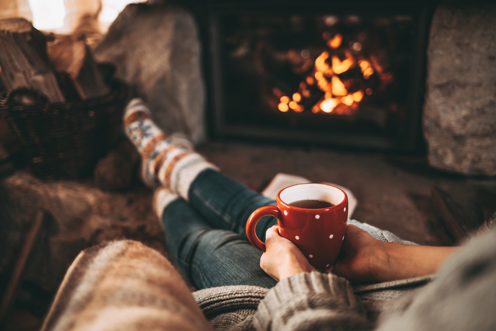 Person sitting indoors in front of a fire with a warm drink in their hands.