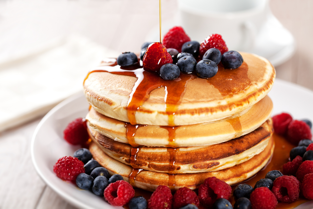Maple syrup being poured on a stack of pancakes topped with berries.
