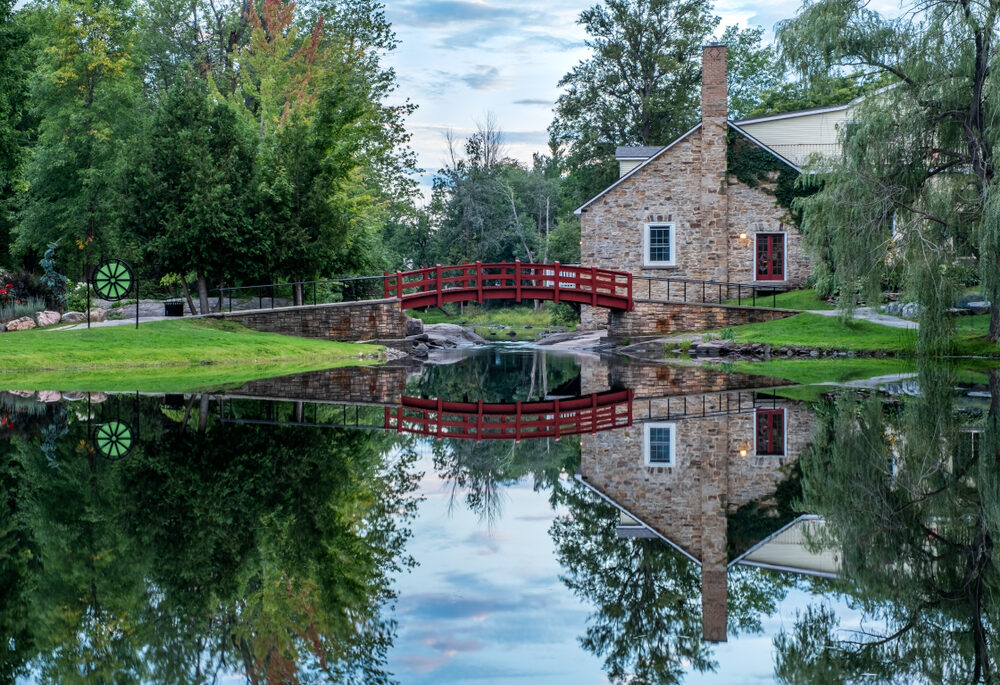 Small stone cottage with a red bridge over a clear river and a tree landscape.