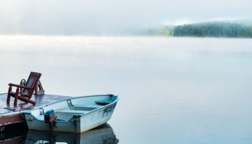 An old boat next to a wooden dock on a still, misty lake.