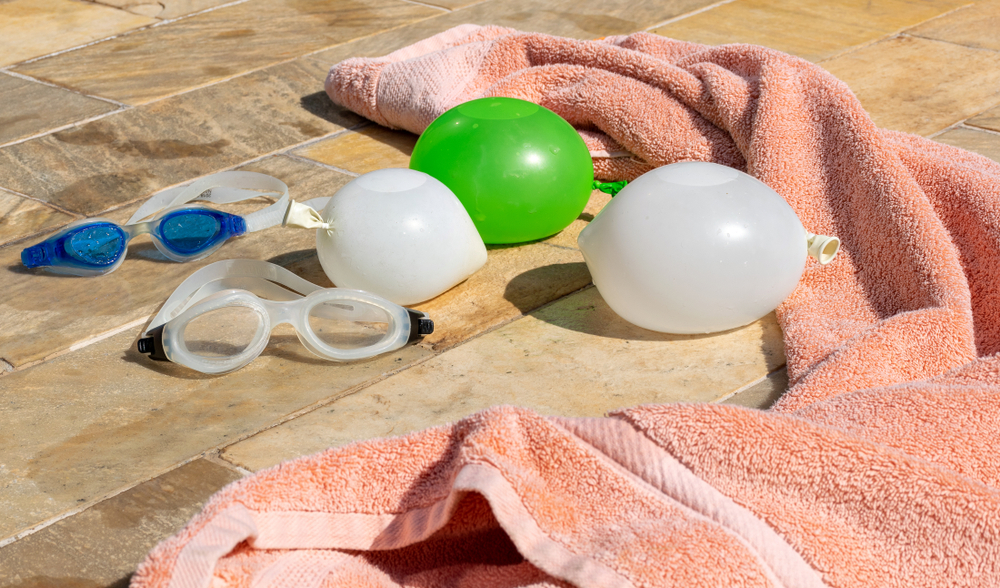 Water balloons and swimming goggles next to a pink beach towel on a summer day.