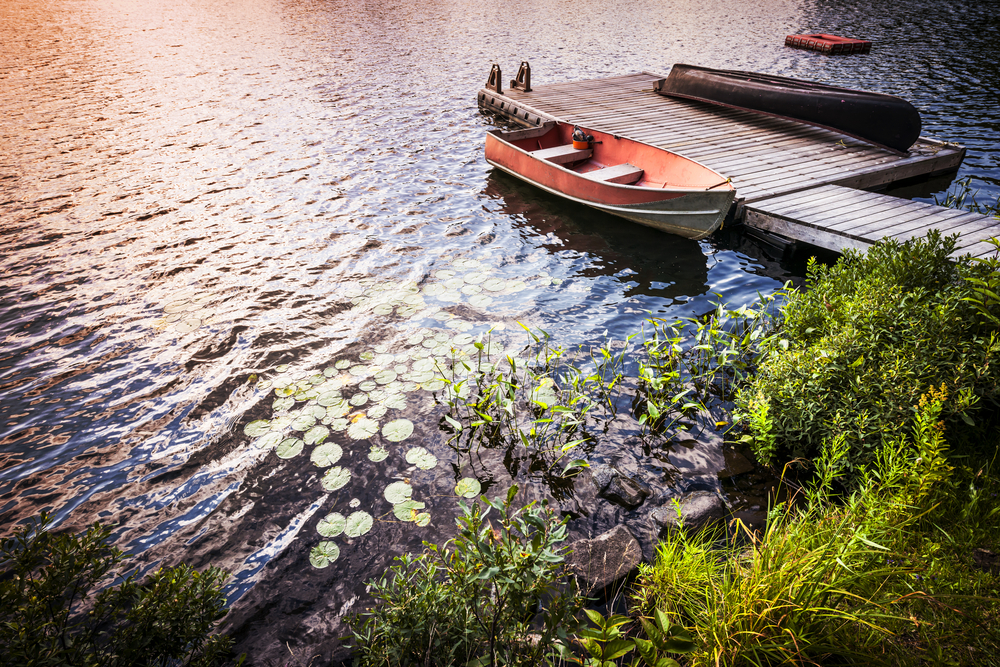 An old red rowboat anchored next to a wooden dock on a lake with the sun reflecting on it.