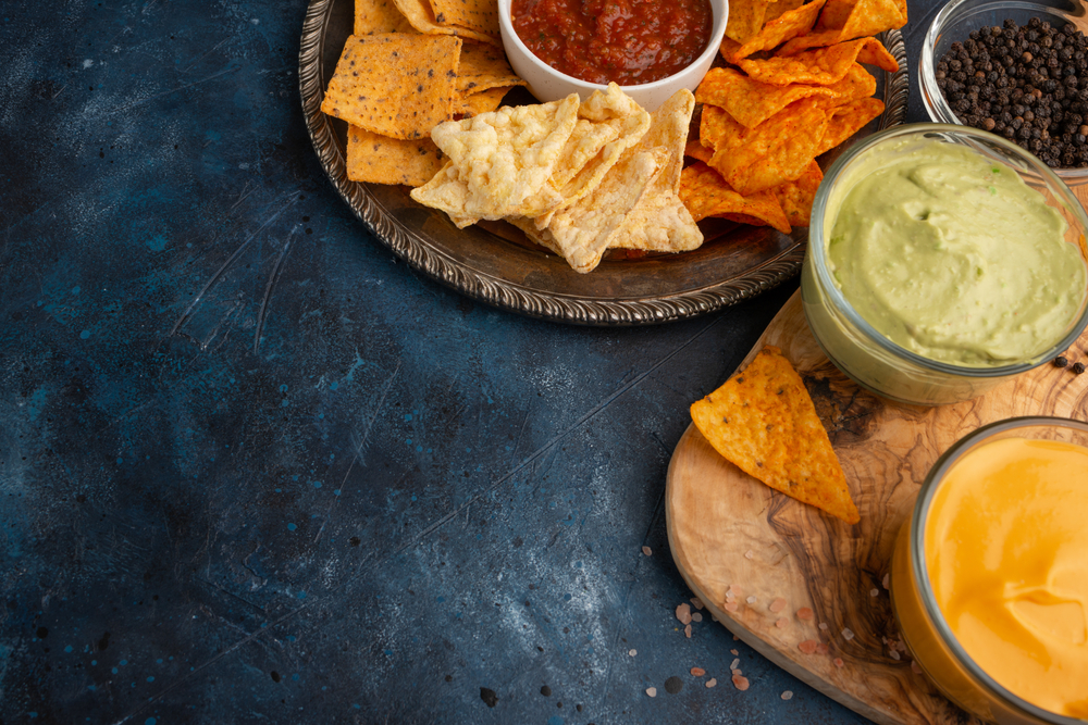Assorted chips with dips on wooden boards against a dark stone background.