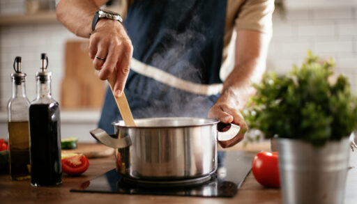 Man in grey apron cooking food in a pot in a kitchen.
