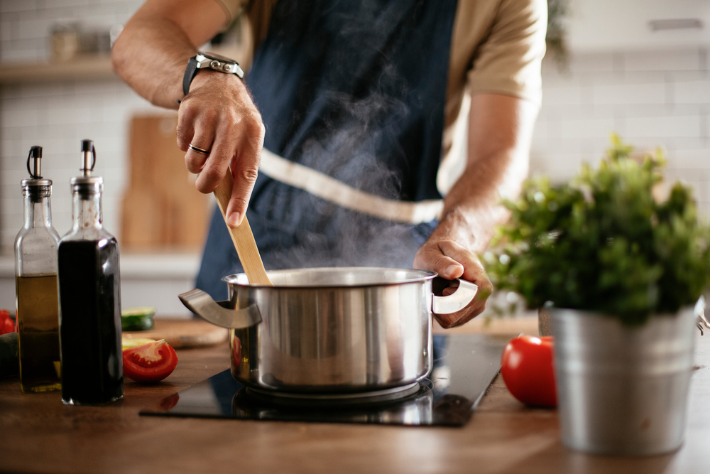 Man in grey apron cooking food in a pot in a kitchen.