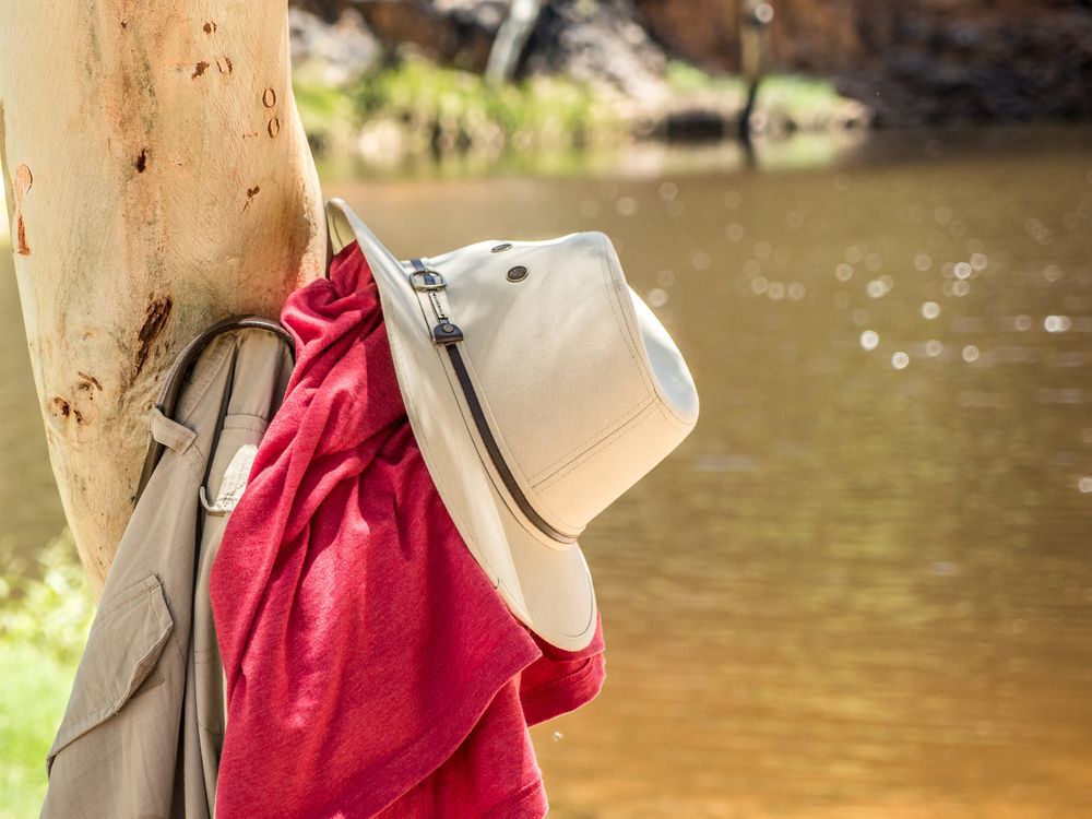 Red shirt and a beige hat and pants hung on a tree next to a river.