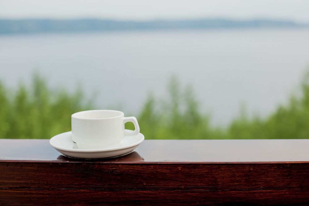 White coffee cup and saucer on a wooden deck railing overlooking a lake.