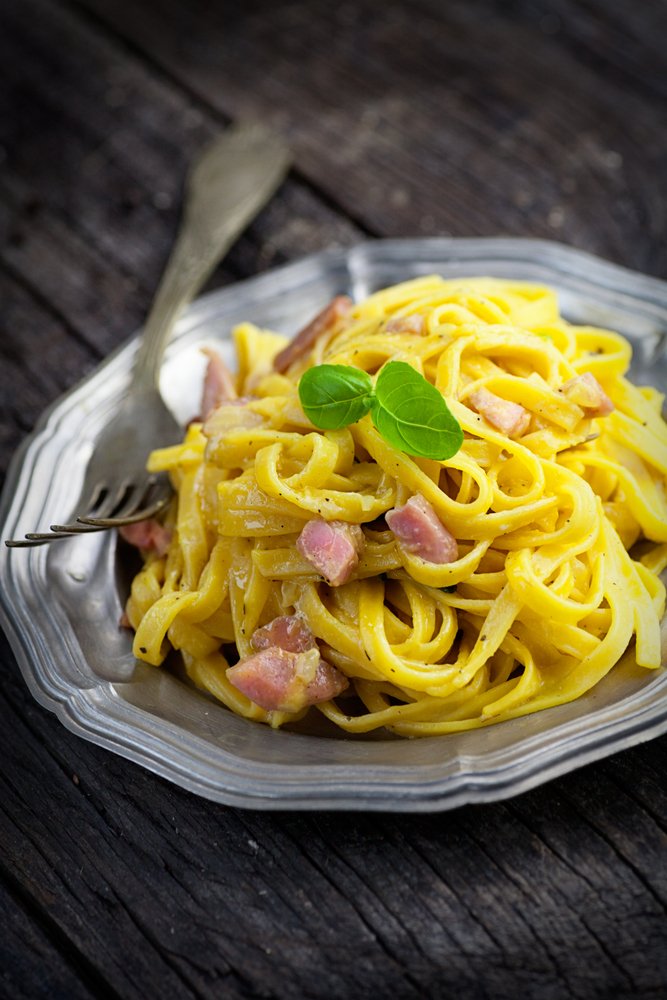 Pasta carbonara with ham and a basil leaf on top on a silver plate.