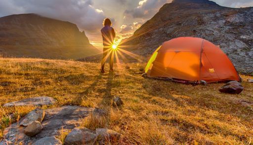 Man standing next to an orange tent on a mountain facing a sunset.
