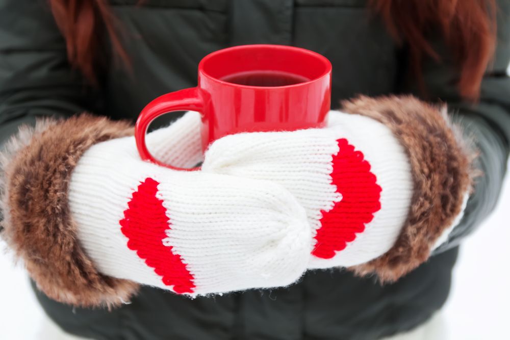 Woman wearing white mittens with read hearts on them holding a red mug of hot cocoa.