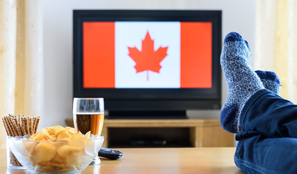 Person with their feet up and a bowl of chips and beer next to them with a Canadian flag displayed on a TV.