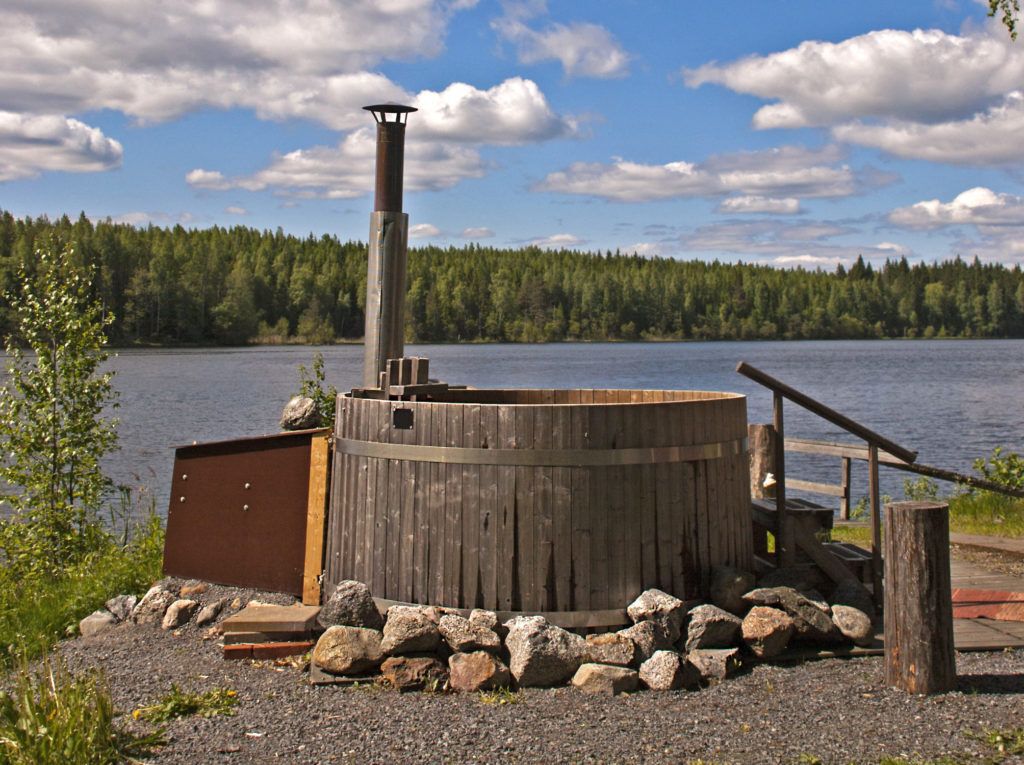 Outdoor wooden hot tub by a lake bordered with a forest in the summer.