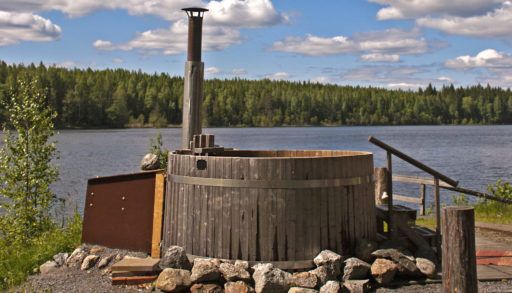 Outdoor wooden hot tub by a lake bordered with a forest in the summer.
