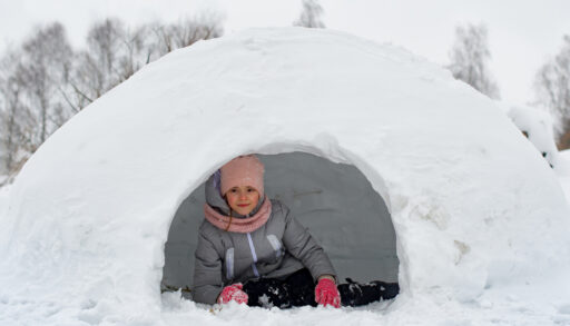 Child in a grey snowsuit laying on the ground in an igloo.