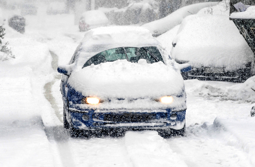 Blue car covered in snow improperly cleared off driving on the road.