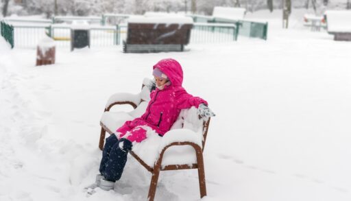 Bored little girl in pink snowsuit sitting on a bench covered in snow.