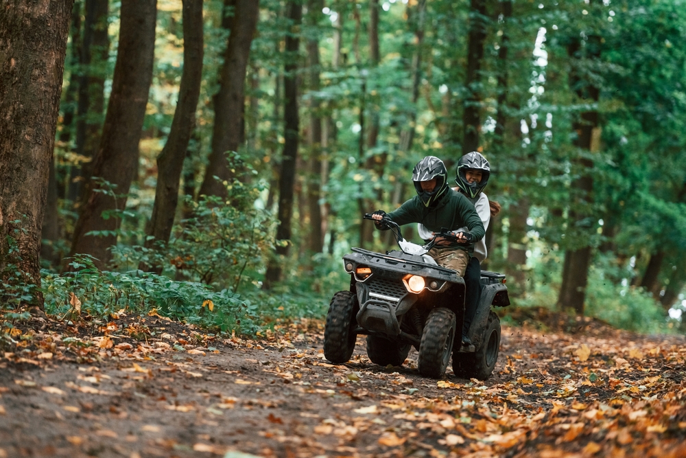 Two people riding an ATV on a trail in a thickly wooded forest.