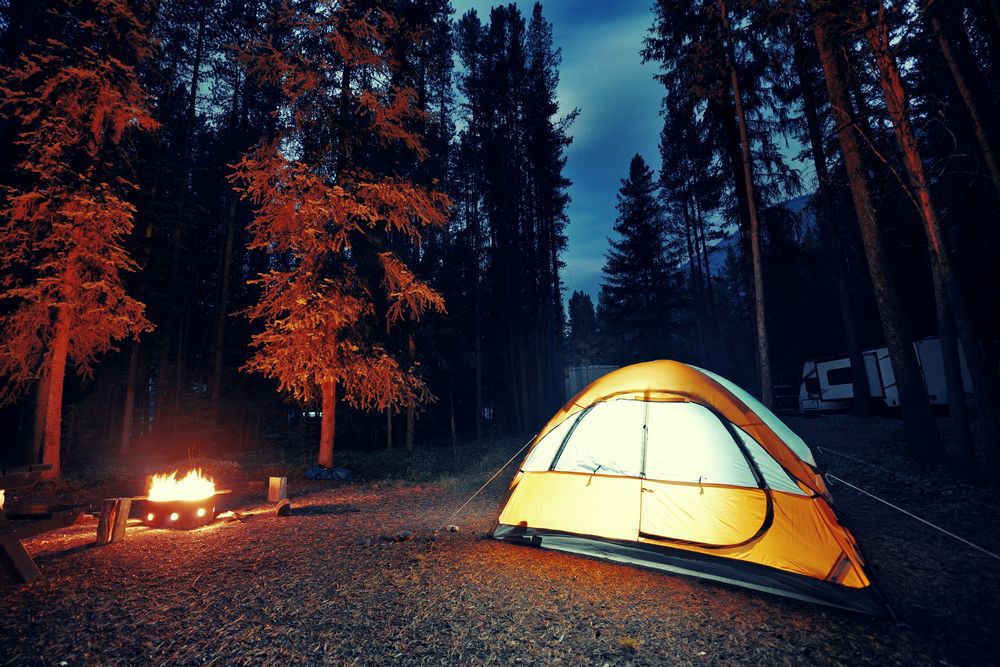 Yellow tent lit up next to a fire at dusk in the woods.