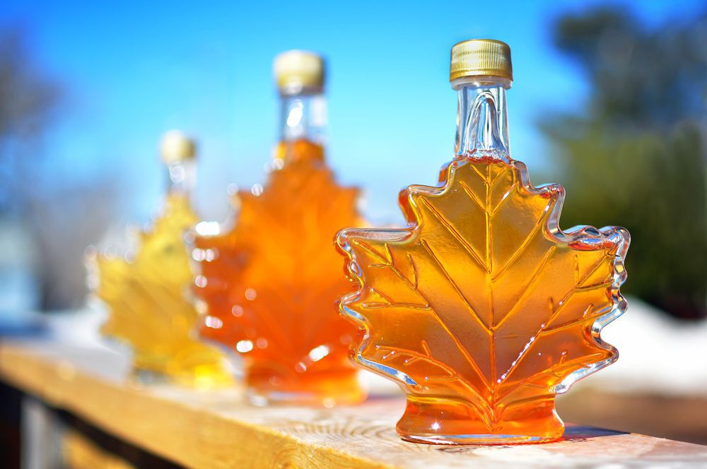 Three maple leaf shaped maple syrup bottles filled with varying shades of maple syrup on an outdoor railing.