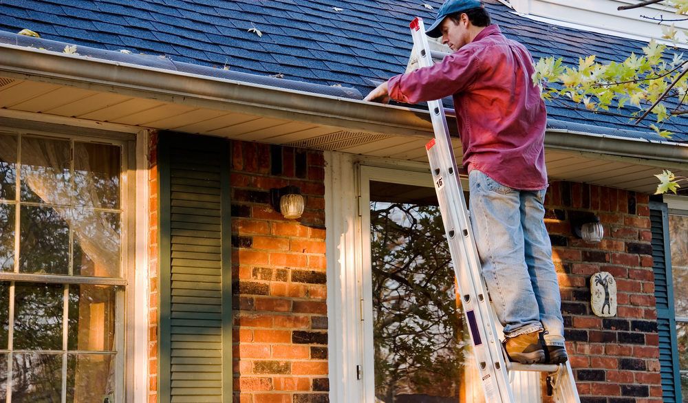 Man in a red plaid shirt and a blue hat on a ladder cleaning out the gutters of his house.