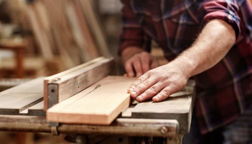 Man in a plaid shirt feeding a piece of wood through a table saw in a workshop.