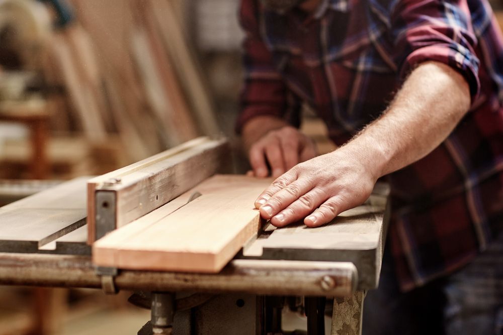 Man in a plaid shirt feeding a piece of wood through a table saw in a workshop.