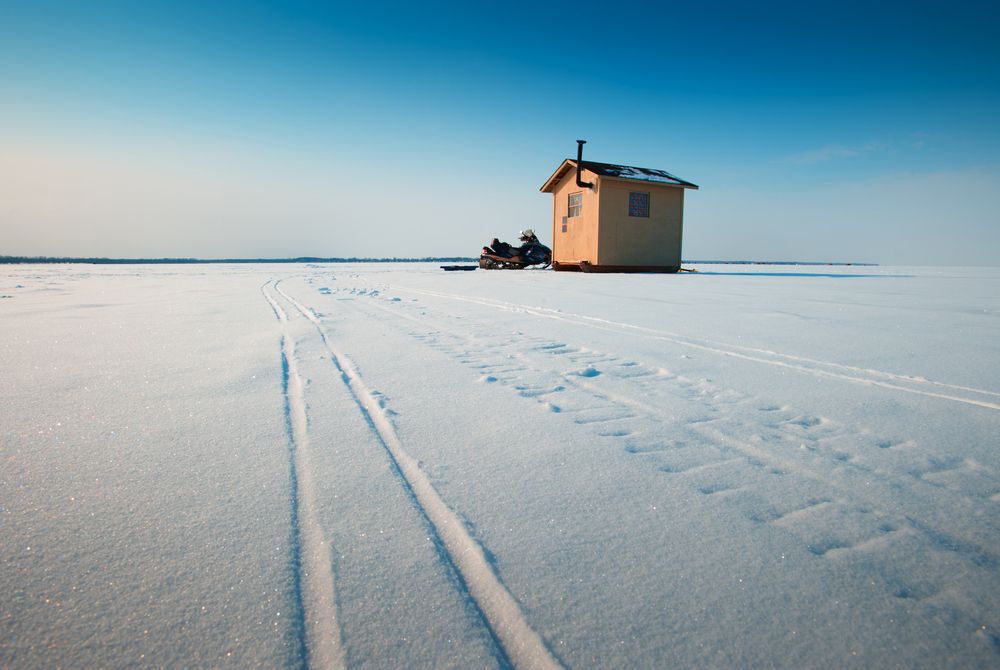 An ice fishing hut sits on a frozen lake with a snowmobile parked outside