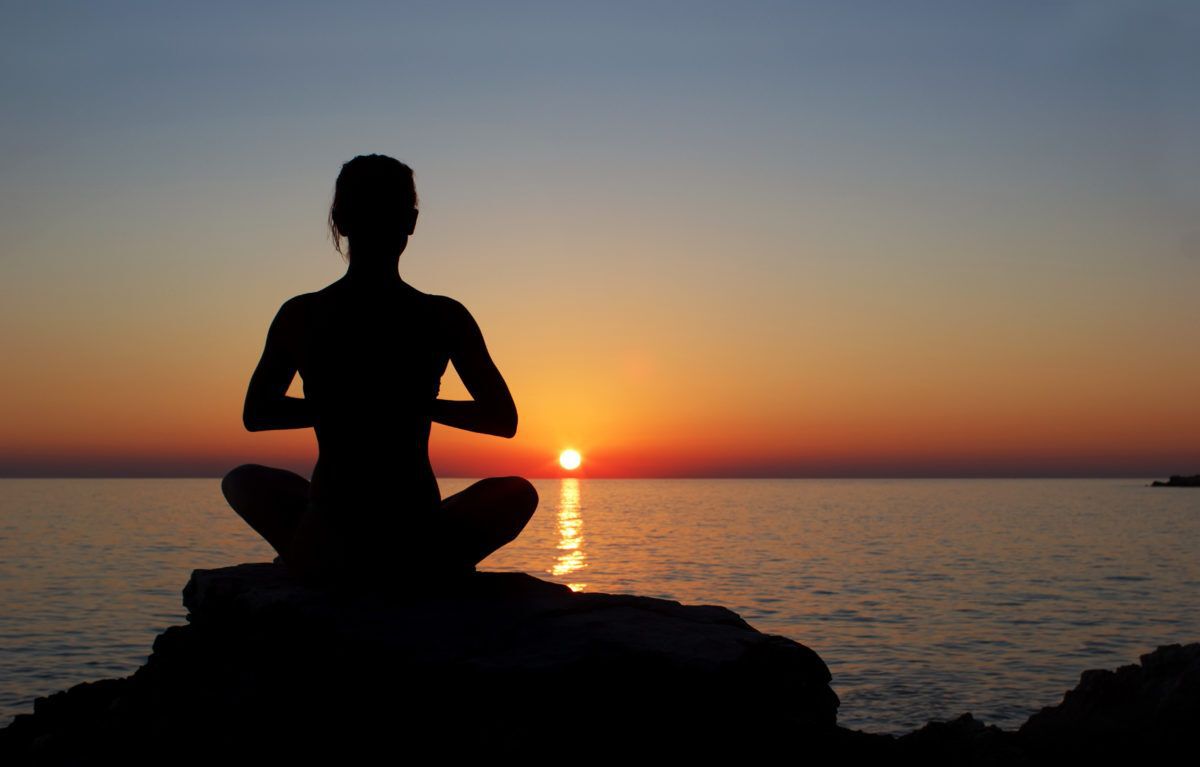 Silhouette of a person practicing meditation on a rock by the ocean at sunset.