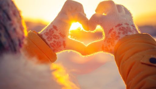 A woman holds mittened hands up in the shape of a heart as the sun sets