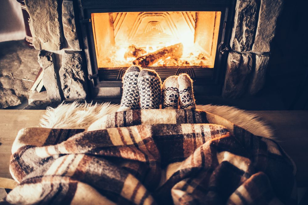 Two sets of feet in socks covered by a warm blanket sit in front of a fire