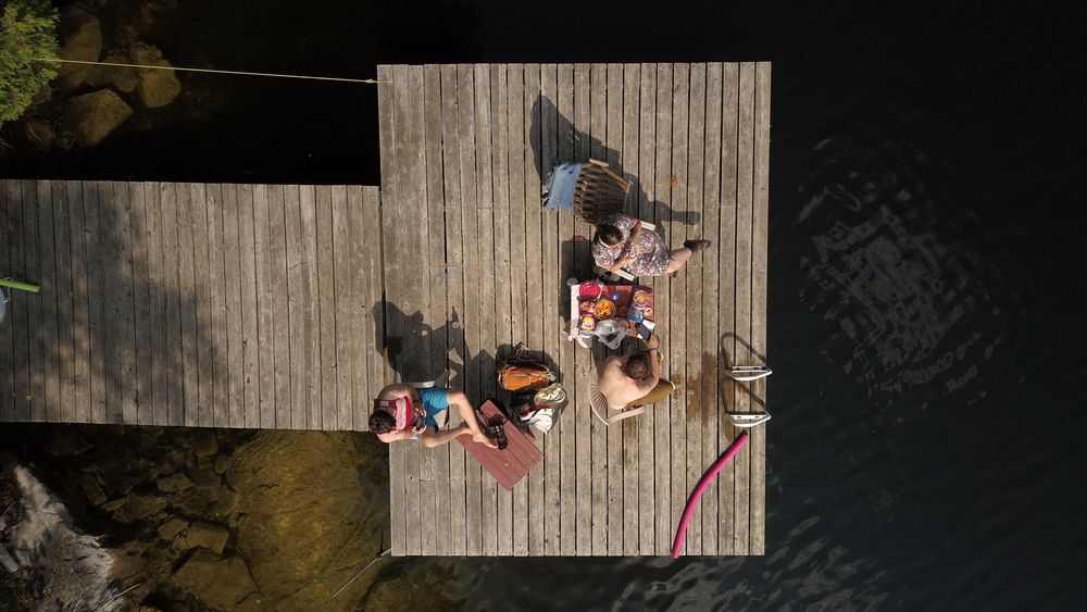 Top down aerial drone image of people sitting on a wooden dock at a cottage lake.