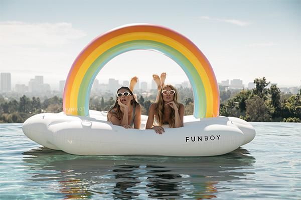 Two women lie on a pool float in the shape of a cloud and rainbow